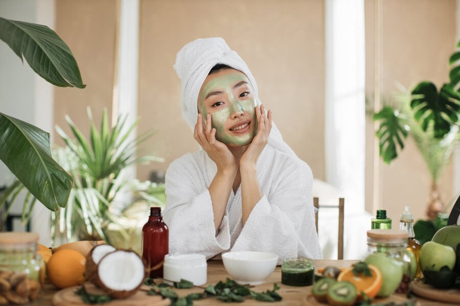 Người ngồi trong spa, đắp mặt nạ xanh, quấn khăn, xung quanh có trái cây và sản phẩm chăm sóc da.