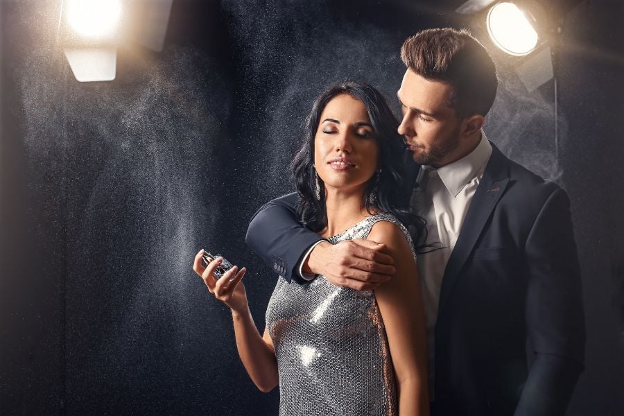 Một người phụ nữ cầm chai nước hoa, đứng gần một người đàn ông mặc vest, ánh sáng chiếu sáng từ phía sau.