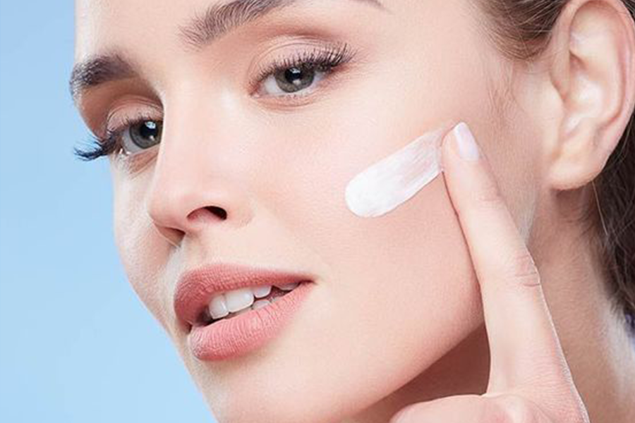 Người phụ nữ thoa kem dưỡng da lên má với nền trời xanh nhạt.