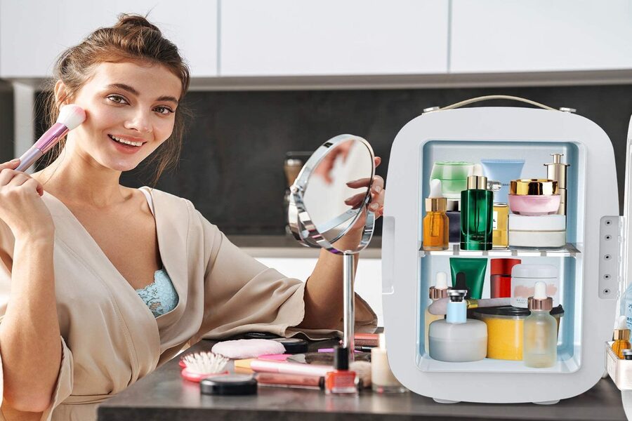 Người phụ nữ trang điểm trước gương, bên cạnh là tủ lạnh nhỏ chứa nhiều sản phẩm chăm sóc da.