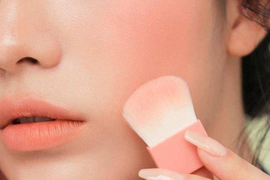 Cận cảnh một người đang dùng cọ trang điểm má hồng, môi và má có màu sắc tự nhiên và hài hòa.
