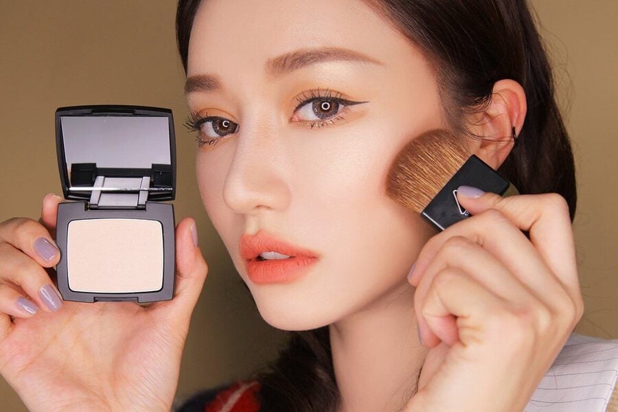 Người đang trang điểm với phấn phủ và cọ, thể hiện cách sử dụng phấn má hồng lên gò má.
