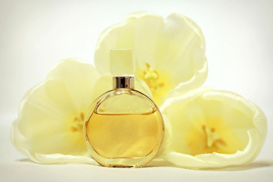 Chai nước hoa tròn, màu vàng, đặt trước ba bông hoa màu trắng nhạt, tạo nền tươi sáng và thanh lịch.