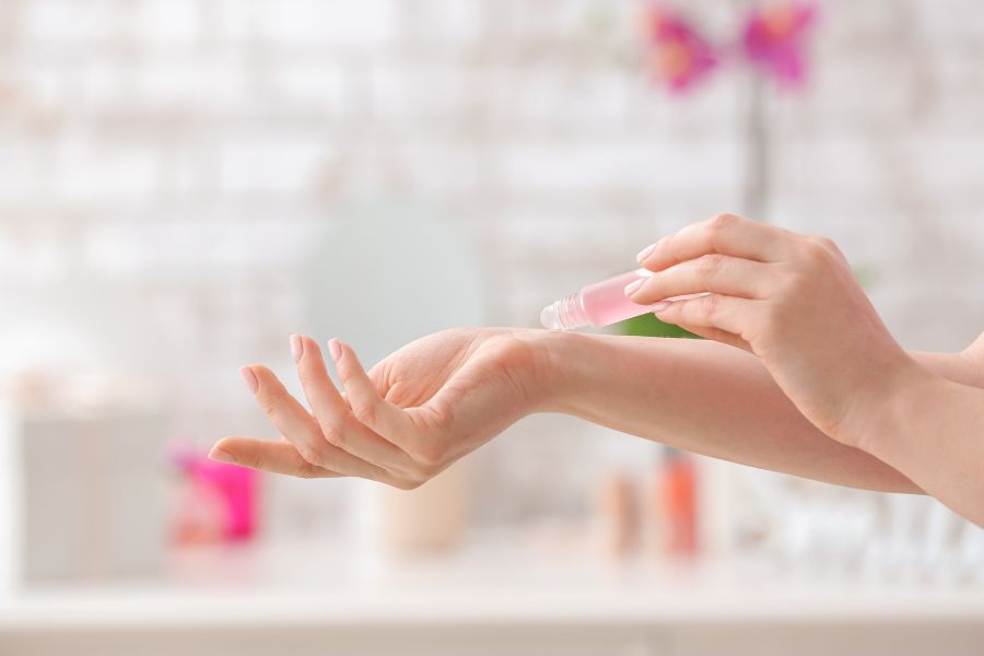 Người đang thoa dầu thơm từ chai nhỏ lên cổ tay, nền mờ với vật dụng trang trí.