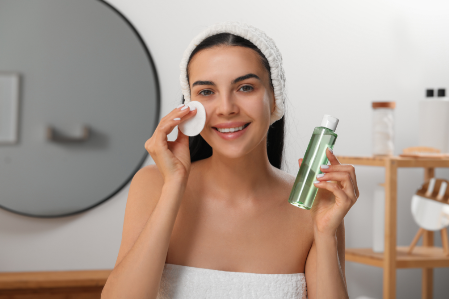 Người phụ nữ đang dùng bông tẩy trang và cầm chai dung dịch dưỡng da trong phòng tắm.