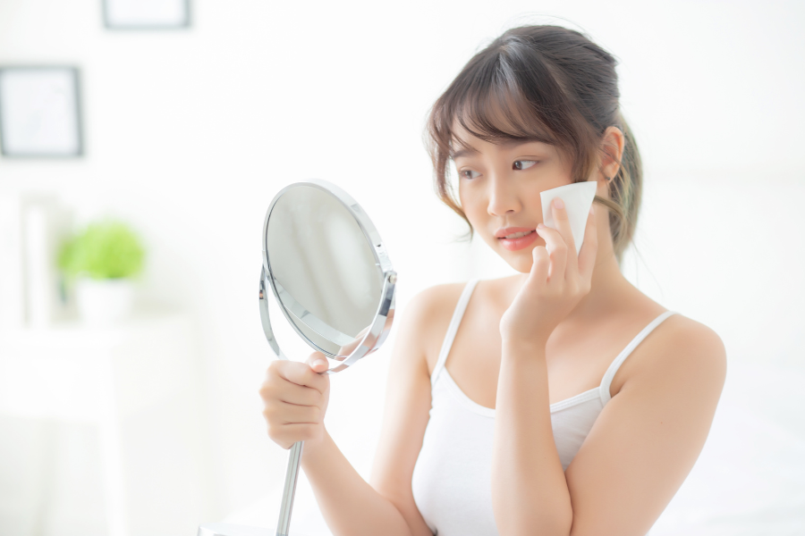 Người đang soi gương và dùng bông tẩy trang, mặc áo ba lỗ trắng trong không gian sáng.