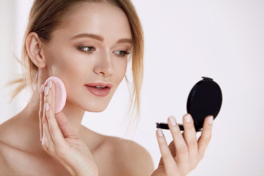 Người đang trang điểm, tay cầm bông phấn và gương nhỏ, nền trắng.