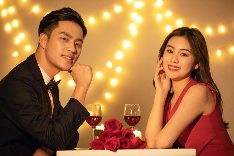 Cặp đôi ngồi tại bàn ăn tối lãng mạn, có hoa hồng và ly rượu vang, ánh đèn vàng lung linh phía sau.