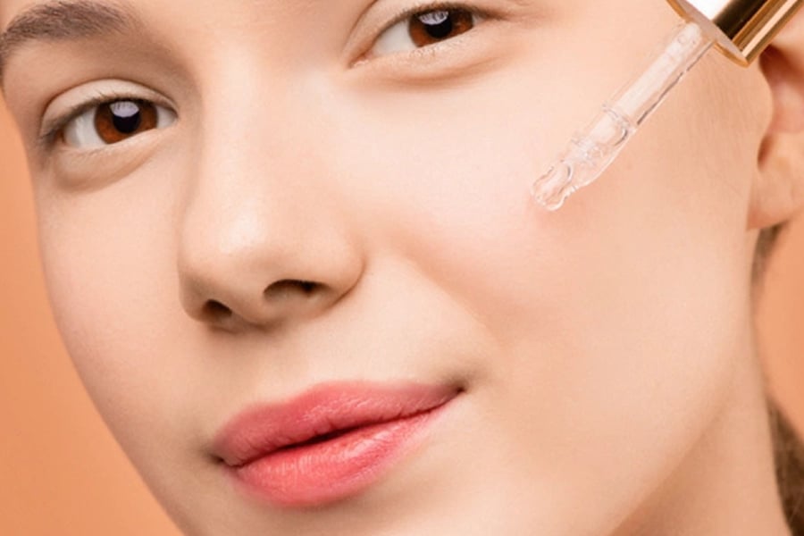 Người đang thoa serum lên má bằng một ống nhỏ giọt trên nền màu cam nhạt.
