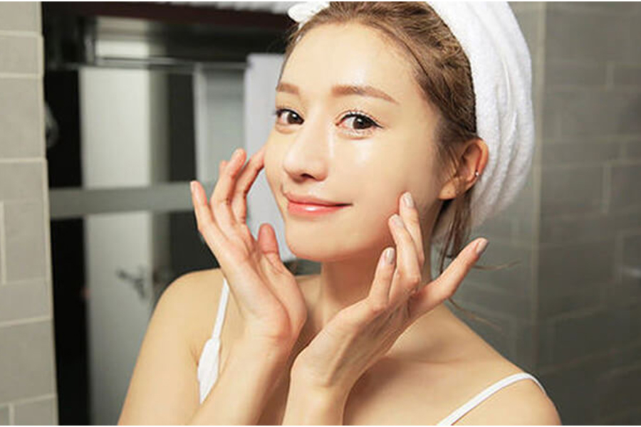 Người phụ nữ với khăn quấn tóc đang chạm vào má, tươi cười trong phòng tắm, ánh sáng tự nhiên chiếu sáng.