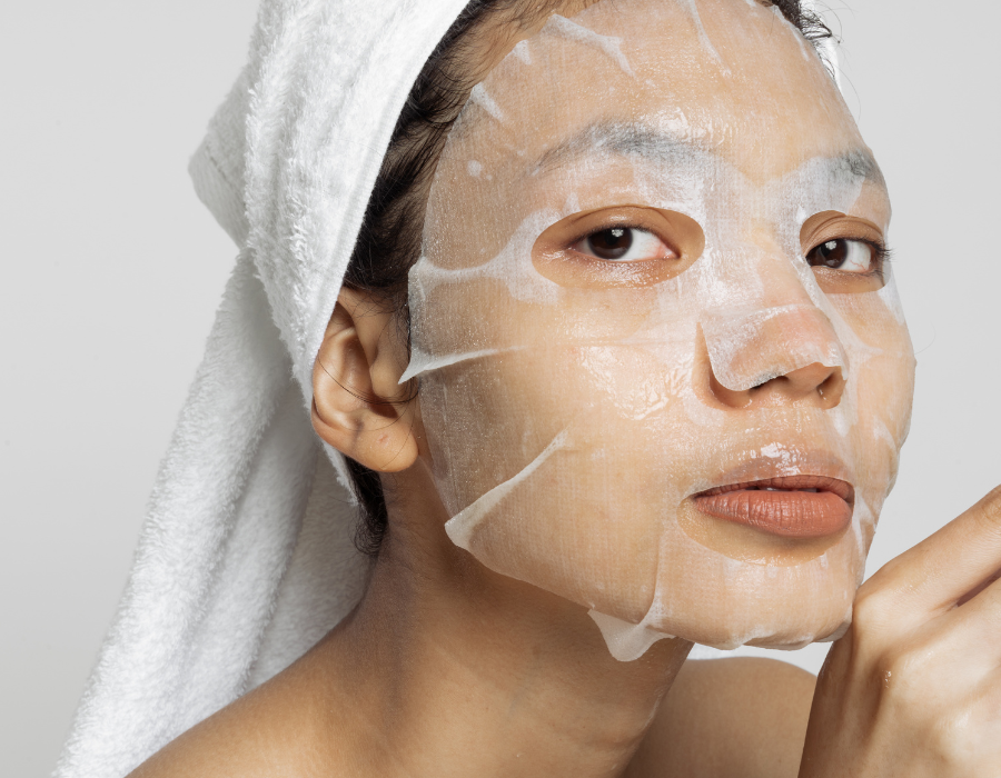 Người đang đắp mặt nạ giấy dưỡng da, quấn khăn trắng trên đầu, nhìn thẳng về phía máy ảnh.