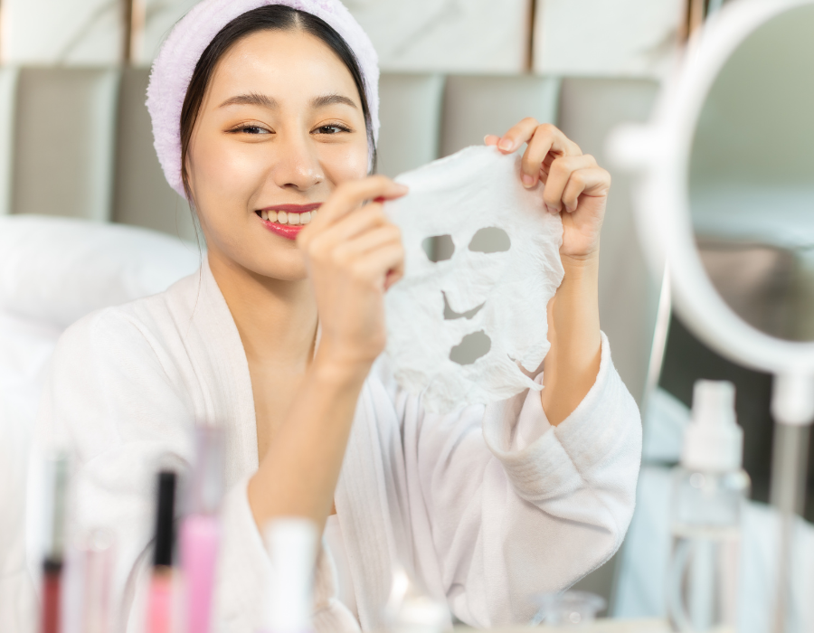 Người cầm mặt nạ giấy dưỡng da, ngồi trước gương với nhiều sản phẩm chăm sóc sắc đẹp xung quanh.