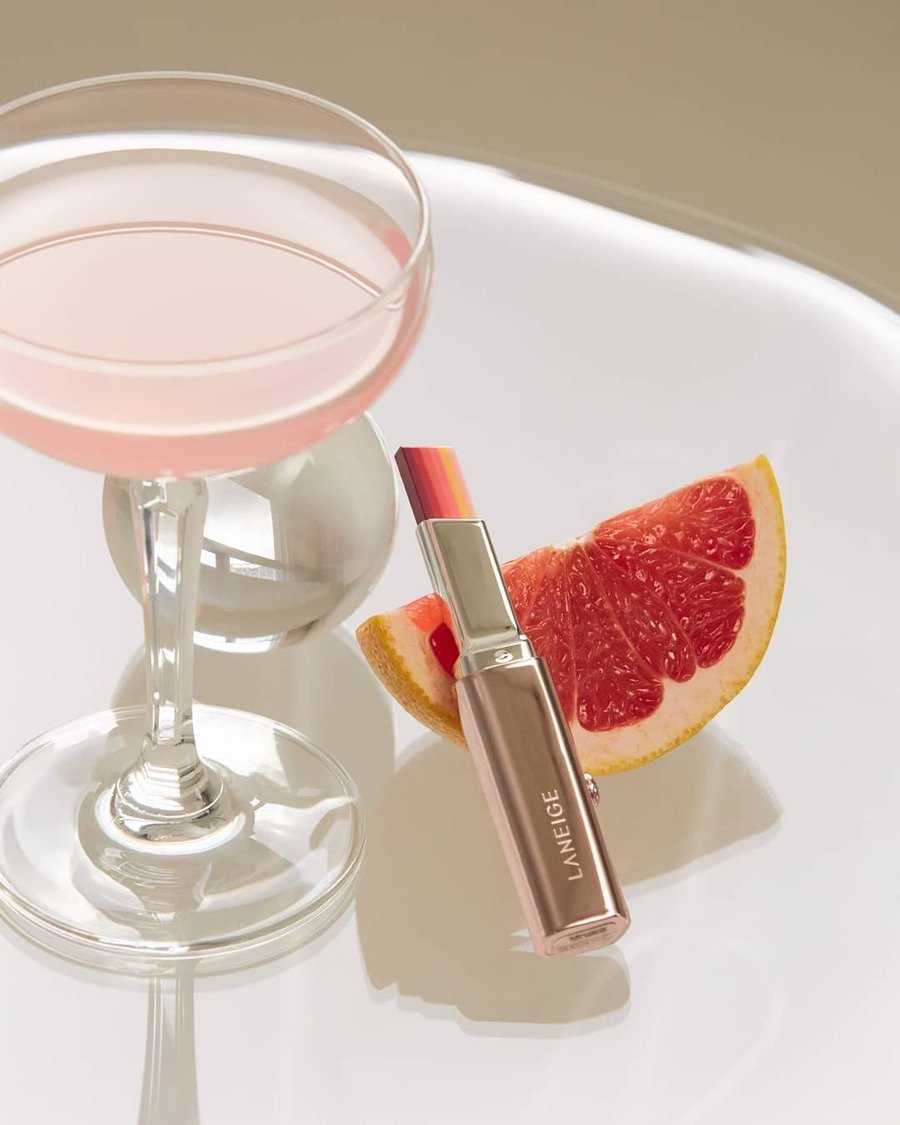 Ly cocktail màu hồng, son môi và lát bưởi đỏ trên bề mặt trắng sáng.