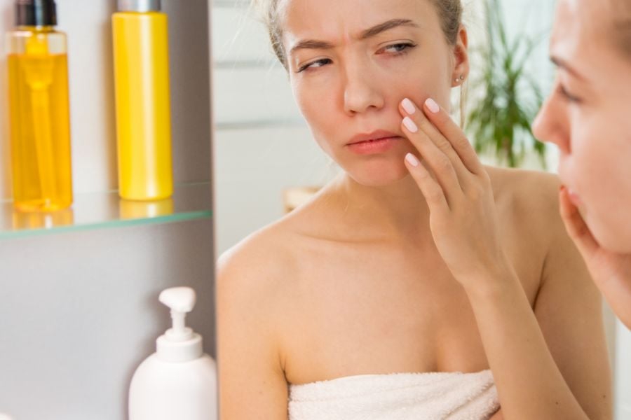 Người phụ nữ nhìn vào gương, chạm vào mặt, biểu hiện lo lắng, có chai lọ mỹ phẩm bên cạnh.