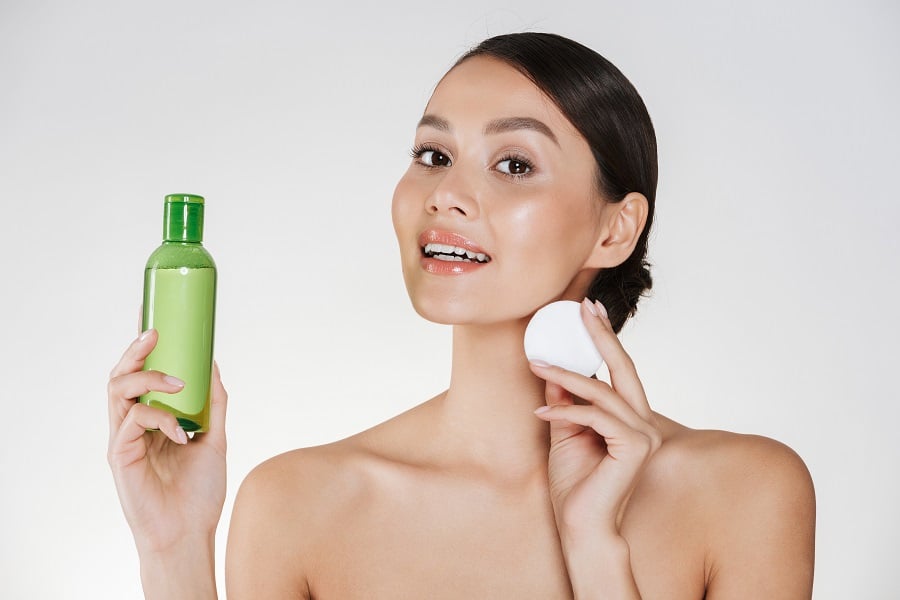 Người phụ nữ cầm chai mỹ phẩm màu xanh và miếng bông tẩy trang trên nền trắng.