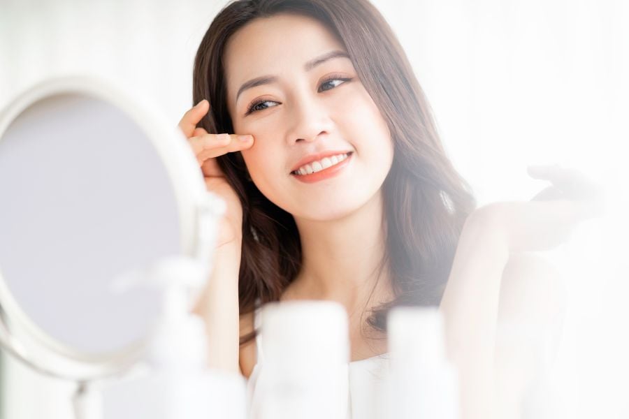 Người phụ nữ đang mỉm cười, nhìn vào gương, sử dụng sản phẩm dưỡng da với chai lọ phía trước.