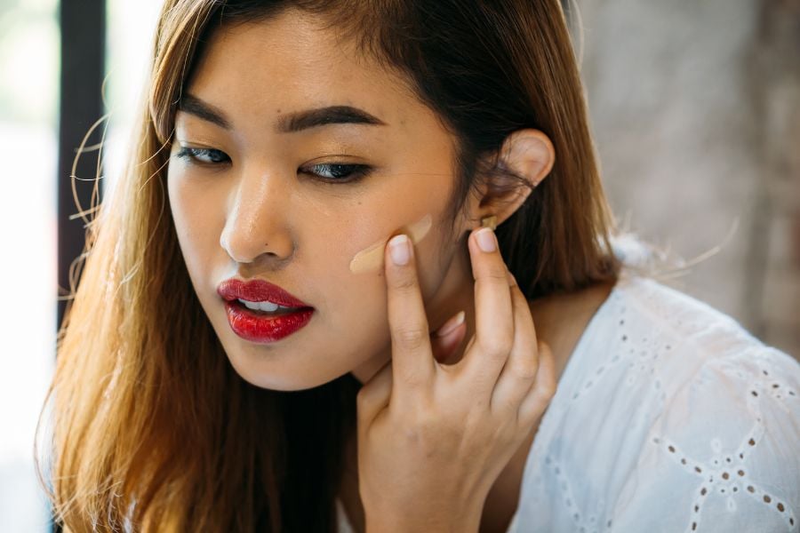Người phụ nữ đang thoa kem nền lên má, mặc áo trắng và có mái tóc dài màu nâu.