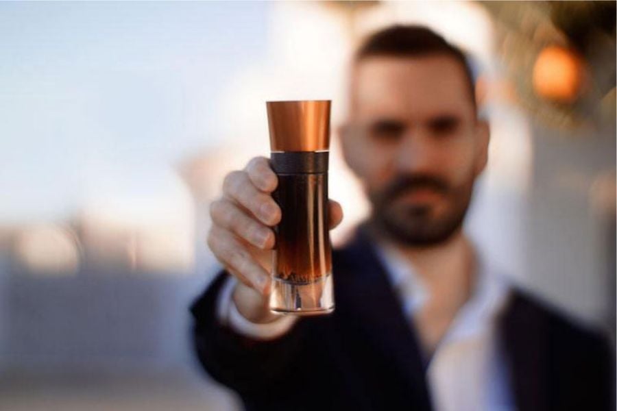 Người đàn ông cầm chai nước hoa, phía sau là phông nền mờ, chai có nắp màu đồng.