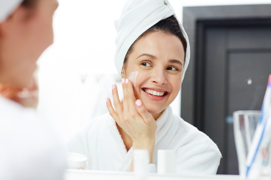 Người phụ nữ trong áo choàng tắm, đội khăn, thoa kem dưỡng lên mặt trước gương, đang mỉm cười.