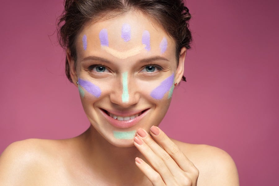 Người phụ nữ cười, mặt có các vệt trang điểm màu tím và xanh, nền hồng.