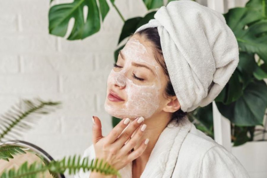 Người đang thư giãn với mặt nạ dưỡng da, quấn khăn trên đầu, nền có cây xanh.