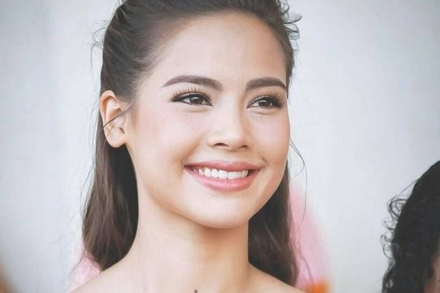 Người phụ nữ tóc dài buộc gọn, đang mỉm cười với phông nền màu sáng.