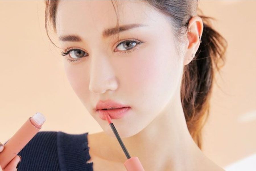 Người phụ nữ đang thoa son môi màu hồng nhạt, mặc áo xanh đậm, nhìn thẳng vào máy ảnh trong nền sáng.