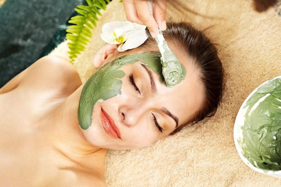 Người đang thư giãn với mặt nạ dưỡng da màu xanh lá trên mặt, có bàn tay đang thoa mặt nạ.