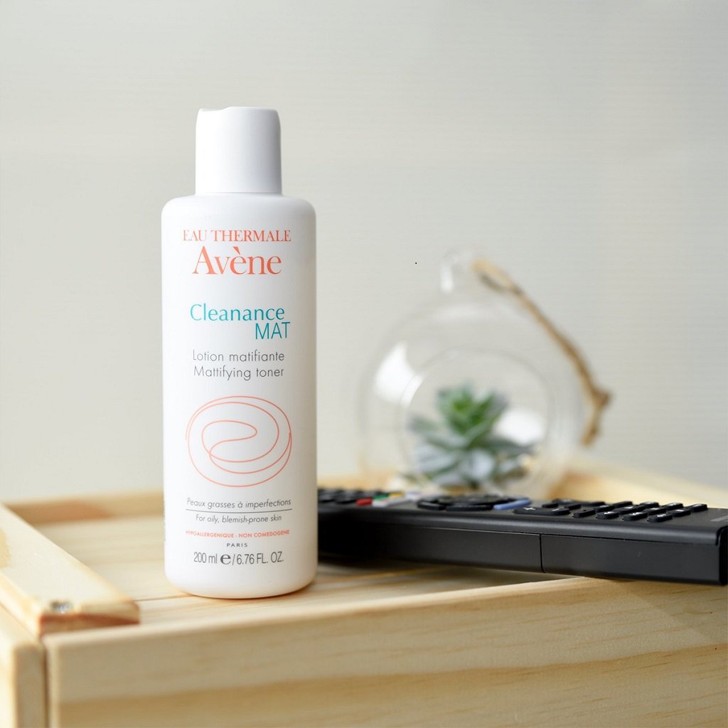 Chai toner đặt trên bề mặt gỗ, bên cạnh là một điều khiển từ xa và một vật trang trí có cây nhỏ bên trong.