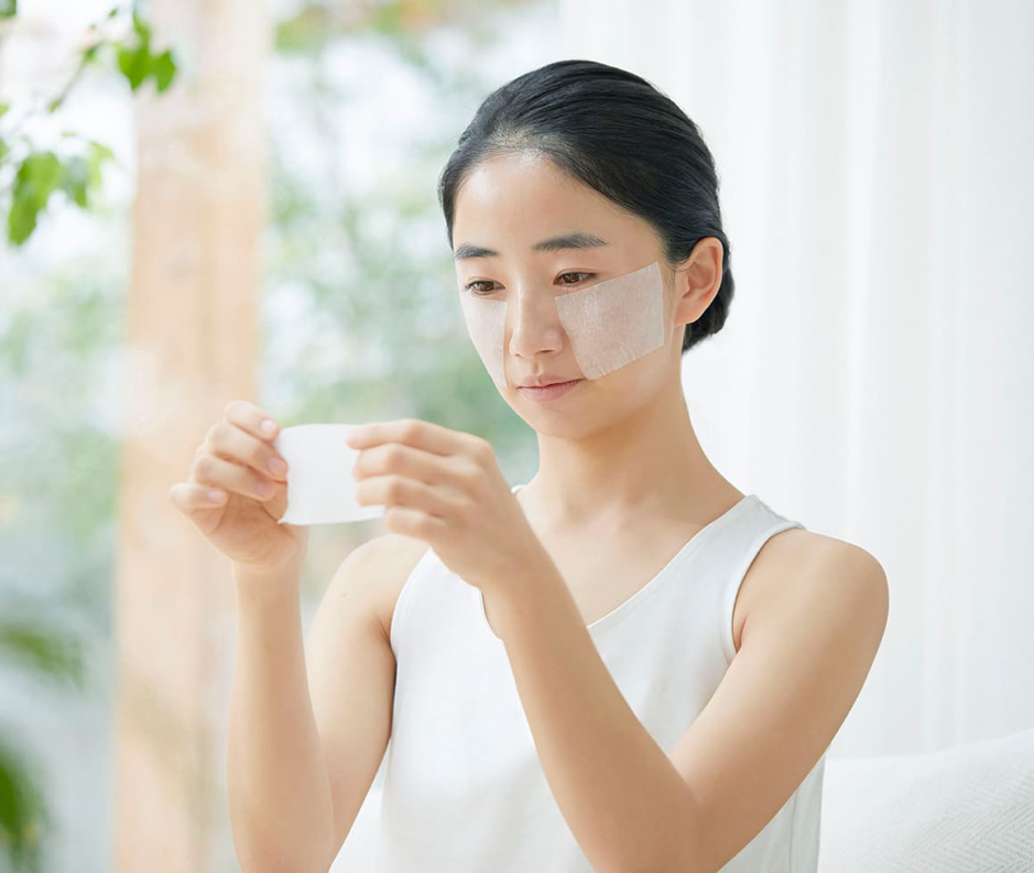 Người phụ nữ ngồi trong nhà, sử dụng mặt nạ dưỡng da. Cô đang cầm một tấm mặt nạ.