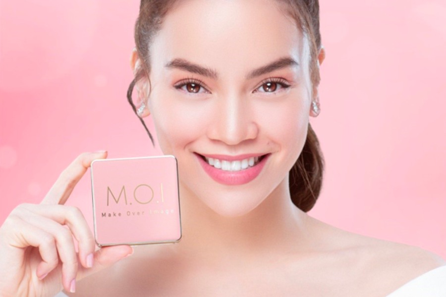 Người phụ nữ với nụ cười tươi cầm hộp mỹ phẩm M.O.I trên nền hồng nhạt.