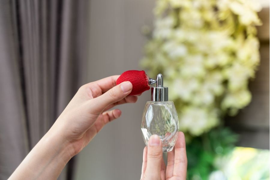 Người đang cầm chai nước hoa thủy tinh với vòi xịt màu đỏ, nền là hoa màu trắng.