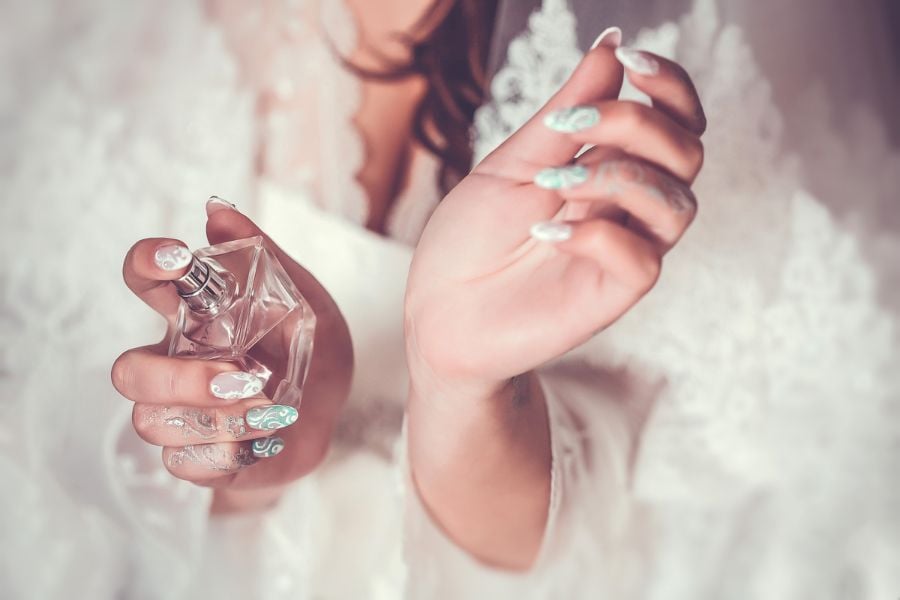 Người với bộ móng tay được trang trí cầm chai nước hoa, nền trắng mờ.